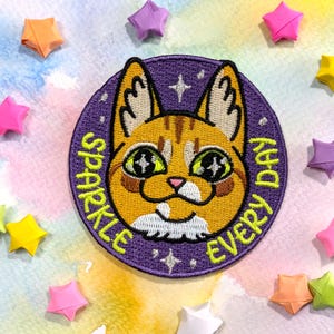 May include: Purple embroidered patch featuring a cartoon orange cat with green eyes. The text "Sparkle Every Day" is written around the cat in a yellow font.