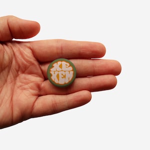 Könnte beinhalten: Ein grüner Knopf mit einem weißen und orangefarbenen Design, auf dem in einer Retro-Schrift "Xem" steht.
