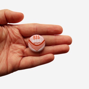 Può includere: Un bottone rosa e arancione con il testo "BIG BELLY BRIGADE" in bianco.