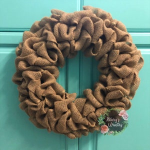 May include: A brown burlap wreath with a ruffled texture. The wreath is hanging on a teal door. The text "Hilary's Wreaths" is visible in the lower right corner of the image.