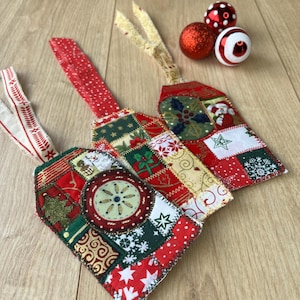 May include: Three fabric gift tags featuring patchwork designs in red, green, gold, and white with holiday motifs like snowflakes, holly, and stars. Each tag has a fabric ribbon loop for hanging, accompanied by three small round ornaments.