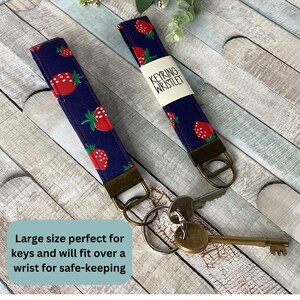 May include: Two navy blue wrist keychains with a red strawberry pattern. Each keychain has a bronze-colored metal ring and a tag that reads "KEYRING WRISTLET". The text "Large size perfect for keys and will fit over a wrist for safe-keeping" is also visible.