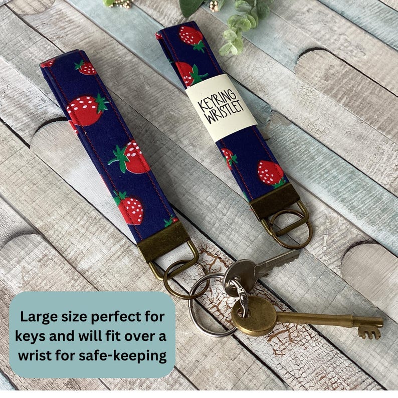 May include: Two navy blue wrist keychains with a red strawberry pattern. Each keychain has a bronze-colored metal ring and a tag that reads "KEYRING WRISTLET". The text "Large size perfect for keys and will fit over a wrist for safe-keeping" is also visible.