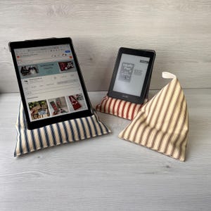 May include: Two fabric tablet and e-reader stands. One stand has a blue and white striped pattern, and the other two have red and white or brown and white striped patterns. A tablet and an e-reader are displayed on the stands.