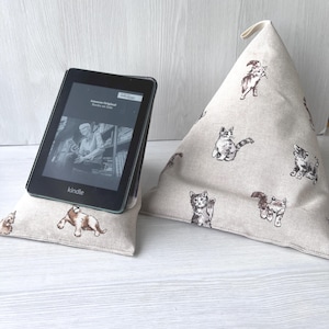 May include: A black Kindle e-reader resting on a beige fabric stand with a repeating cat pattern. The e-reader screen displays text and a black and white image of a person reading. The stand is pyramid-shaped with a small fabric loop at the top.