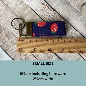 May include: A small, navy blue fabric keychain wristlet with a repeating pattern of red strawberries with white seeds. The keychain includes a bronze-colored metal clasp and ring. The wristlet is 3.3 inches long including hardware and 1 inch wide.