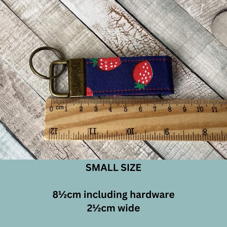 May include: A small, navy blue fabric keychain wristlet with a repeating pattern of red strawberries with white seeds. The keychain includes a bronze-colored metal clasp and ring. The wristlet is 3.3 inches long including hardware and 1 inch wide.