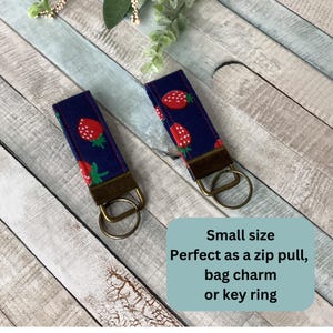May include: Two navy blue key fobs with a repeating pattern of red strawberries and green leaves. Each fob has a bronze-colored metal clasp and key ring. The text on the image says "Small size, Perfect as a zip pull, bag charm or key ring."