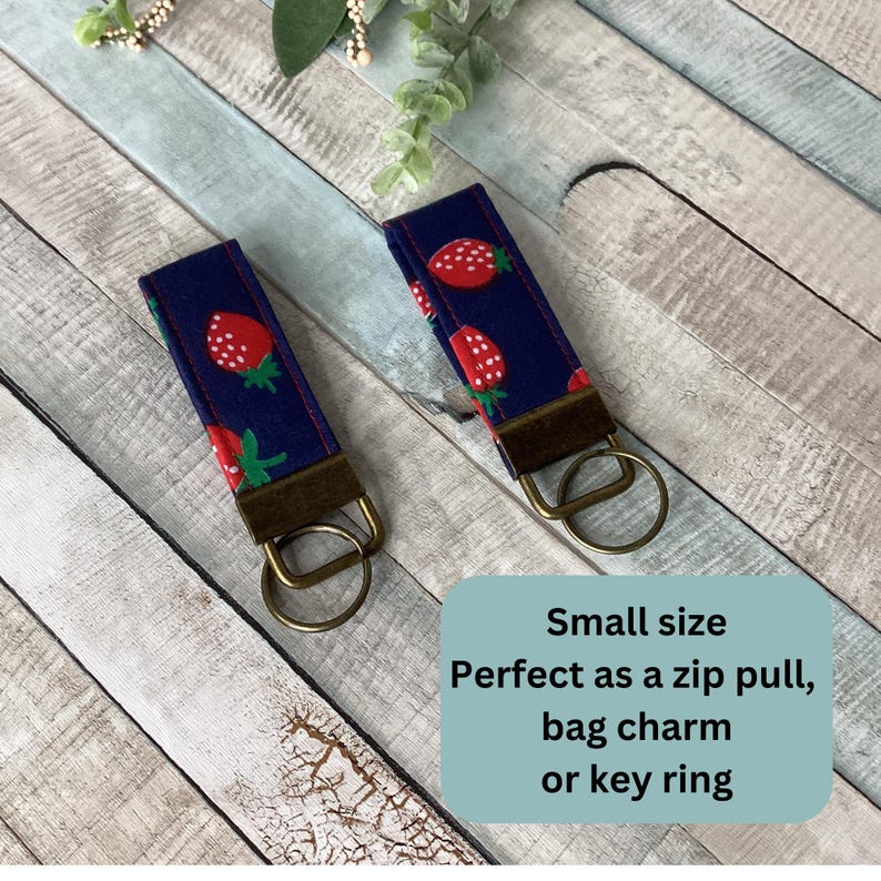 May include: Two navy blue key fobs with a repeating pattern of red strawberries and green leaves. Each fob has a bronze-colored metal clasp and key ring. The text on the image says "Small size, Perfect as a zip pull, bag charm or key ring."