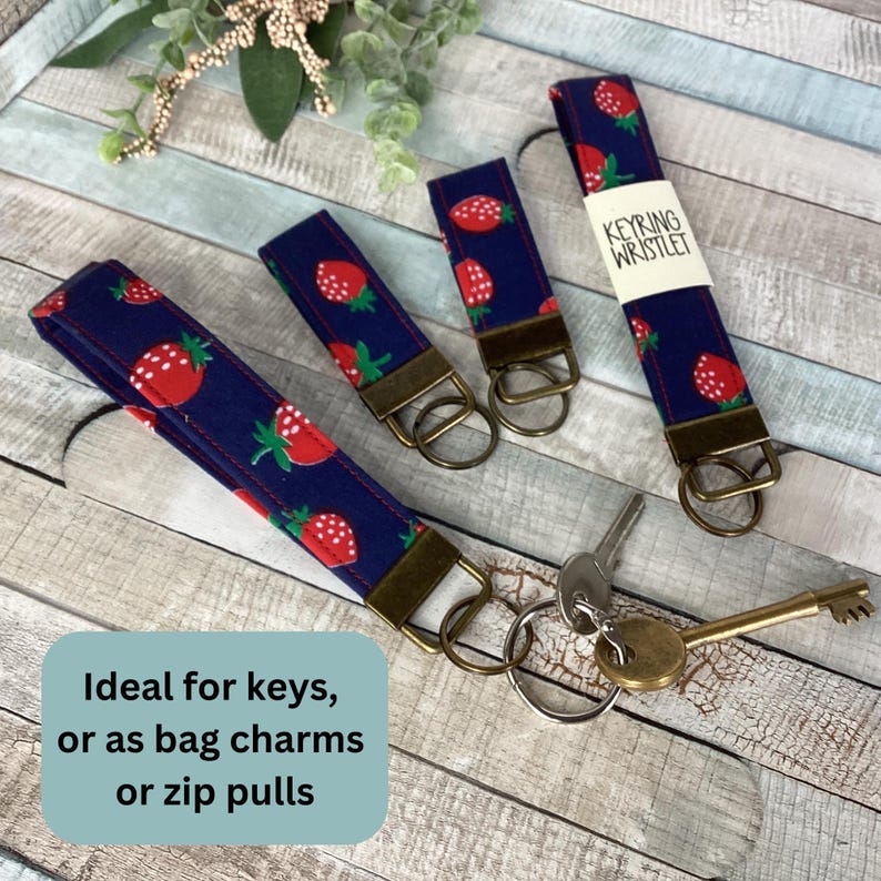 May include: Keyring wristlets with a navy blue background and a pattern of red strawberries with green leaves. The wristlets have a bronze-colored metal ring and are designed to hold keys. One wristlet has a white label that says "KEYRING WRISTLET".