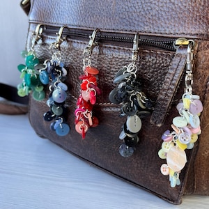 May include: Four handmade button keychains in various colours hang from a brown leather bag. The keychains feature clusters of colourful buttons in shades of green, blue, red, black, and pastel hues, attached to silver-tone metal clasps.