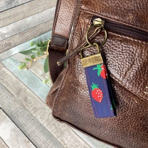 May include: A navy blue fabric keychain with a repeating pattern of red strawberries with green leaves. The keychain is attached to a brown leather bag with a gold zipper and hardware.