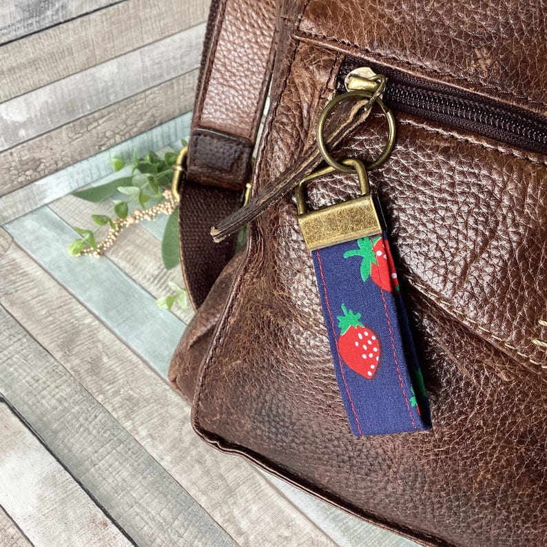 May include: A navy blue fabric keychain with a repeating pattern of red strawberries with green leaves. The keychain is attached to a brown leather bag with a gold zipper and hardware.