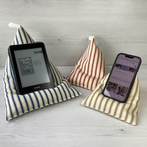 May include: Three triangular fabric stands. One supports a Kindle e-reader, another a smartphone, and the third is empty. The stands feature striped patterns in blue and white, red and white, and beige and white.