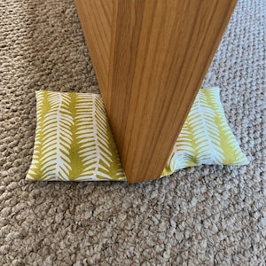May include: A rectangular doorstop featuring a fabric cover with a repeating white fern leaf pattern on a chartreuse green background. The doorstop is positioned against the base of a light brown wooden furniture leg to hold it in place.