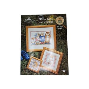 May include: A DMC Collection book titled "Oliver Otter and Friends" with three framed cross-stitch samplers. The samplers feature otters in various outfits. The book is on a green grassy surface.