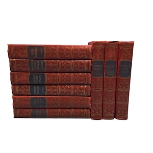 May include: A collection of antique books with red covers and gold embossed designs. The titles include "Robinson Crusoe," "Grimm's Fairy Tales," and "Heidi." The books are stacked and arranged on a white background, showcasing their classic aesthetic.