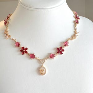 May include: A gold-toned necklace featuring red flower and oval-shaped gemstone accents. The necklace has a pendant with a heart and a small, detailed design. The necklace is displayed on a white mannequin.