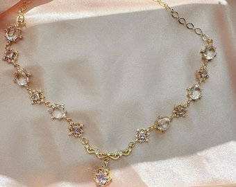 May include: A gold-tone necklace featuring a delicate chain with alternating round and square-cut clear and light purple gemstones. The center of the necklace showcases an ornate filigree pendant drop set with a single light purple stone.