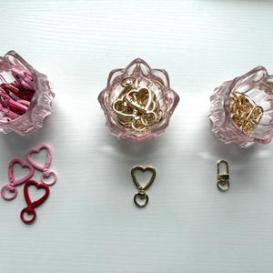 May include: Three light pink glass bowls with scalloped edges containing metal clasps. The left bowl holds pink and red heart-shaped clasps, the center holds gold heart-shaped clasps, and the right holds gold rectangular clasps with loose pieces below.