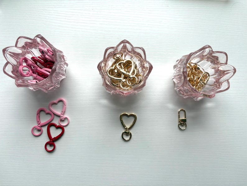 May include: Three light pink glass bowls with scalloped edges containing metal clasps. The left bowl holds pink and red heart-shaped clasps, the center holds gold heart-shaped clasps, and the right holds gold rectangular clasps with loose pieces below.