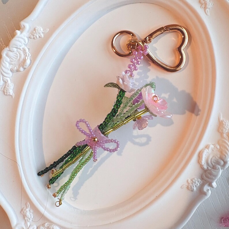 May include: A keychain featuring a bouquet of beaded flowers in shades of pink, white, and green, tied with a small purple beaded bow. The stems are made of green and gold beads, and the entire piece is attached to a gold-toned heart-shaped clasp.