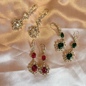 May include: Four pairs of gold dangle earrings with various colored gemstones. The earrings feature intricate designs with small clear rhinestones. The gemstones are red, green, and clear.