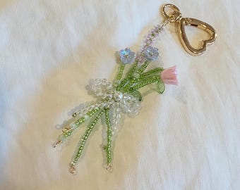 May include: A beaded floral keychain featuring a bouquet of flowers in shades of pink, green, and lavender. The bouquet is accented with clear beads and a pearl, attached to a gold-colored heart-shaped clasp.