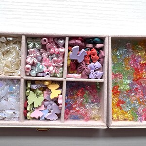 May include: A rectangular jewelry organizer box with multiple compartments filled with an assortment of small bow-shaped charms and beads in various colors, including pearlescent white, pastel pink, blue, yellow, and translucent neon shades.