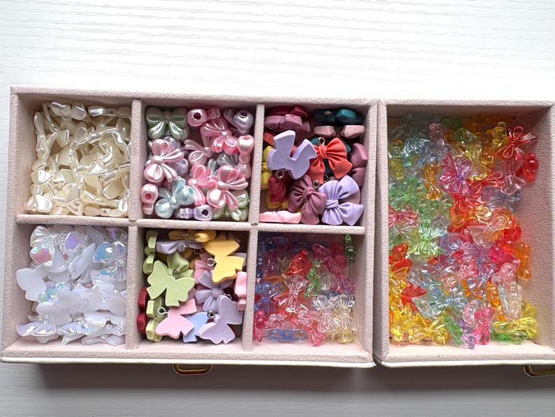 May include: A rectangular jewelry organizer box with multiple compartments filled with an assortment of small bow-shaped charms and beads in various colors, including pearlescent white, pastel pink, blue, yellow, and translucent neon shades.