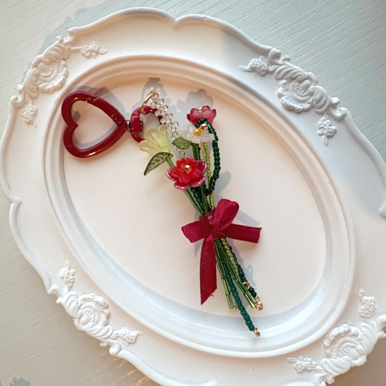 May include: A keychain featuring a red heart-shaped clasp attached to a small bouquet of beaded flowers. The bouquet includes red, white, and pale yellow floral beads with green beaded stems, tied together with a small red fabric ribbon bow.