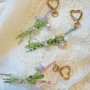 May include: Three beaded floral keychains featuring gold-tone heart-shaped lobster clasps. Each keychain is crafted with green seed beads forming stems and leaves, accented with small pastel-colored flower beads in shades of pink, blue, and white.