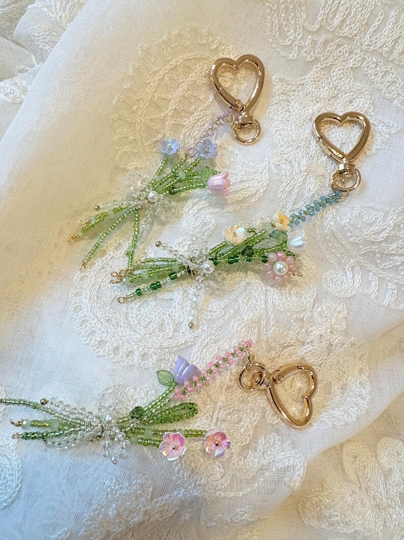 May include: Three beaded floral keychains featuring gold-tone heart-shaped lobster clasps. Each keychain is crafted with green seed beads forming stems and leaves, accented with small pastel-colored flower beads in shades of pink, blue, and white.