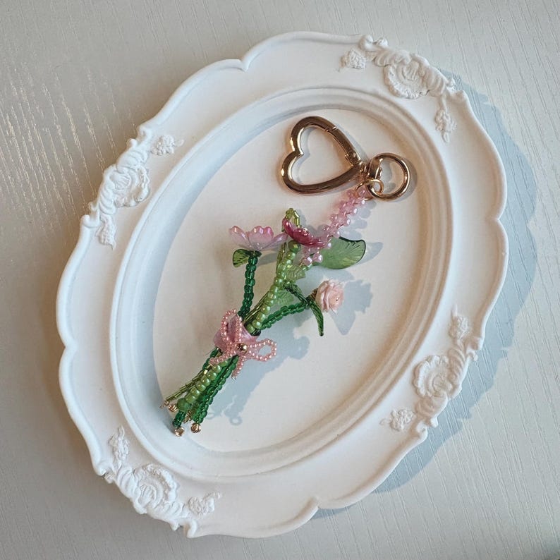 May include: A beaded keychain featuring a bouquet of small pink and green flowers crafted from glass beads and translucent petals. The floral arrangement is tied with a delicate pink beaded bow and attached to a gold-toned heart-shaped metal clasp.