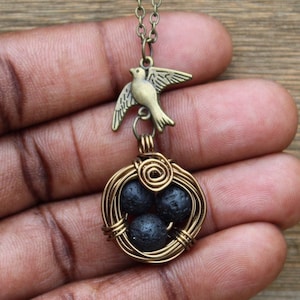 Puede incluir: Un collar de oro con un dije de pájaro y un nido envuelto en alambre con tres cuentas de lava negra.