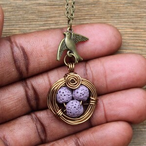 Puede incluir: Un delicado collar con un dije de pájaro y un nido envuelto en alambre con tres cuentas de lava de color lavanda.