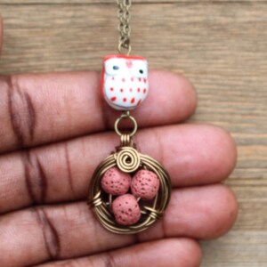 Puede incluir: Un amuleto de búho rojo y blanco cuelga de una cadena dorada, con un nido de alambre que contiene tres huevos rosas debajo.