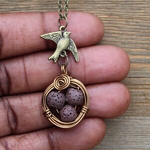 Puede incluir: Un collar de bronce con un dije de pájaro y un colgante envuelto en alambre que contiene cuentas de lava marrón. El colgante se asemeja a un nido de pájaro.