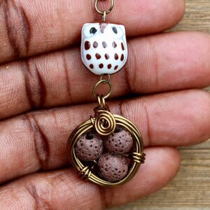 Puede incluir: Un collar de cadena dorada con un dije de búho marrón y blanco y una jaula de alambre con cuentas de lava marrón.