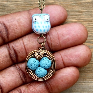 Puede incluir: Un collar con un dije de búho azul y blanco y un dije de nido envuelto en alambre que contiene cuatro cuentas de lava azul.