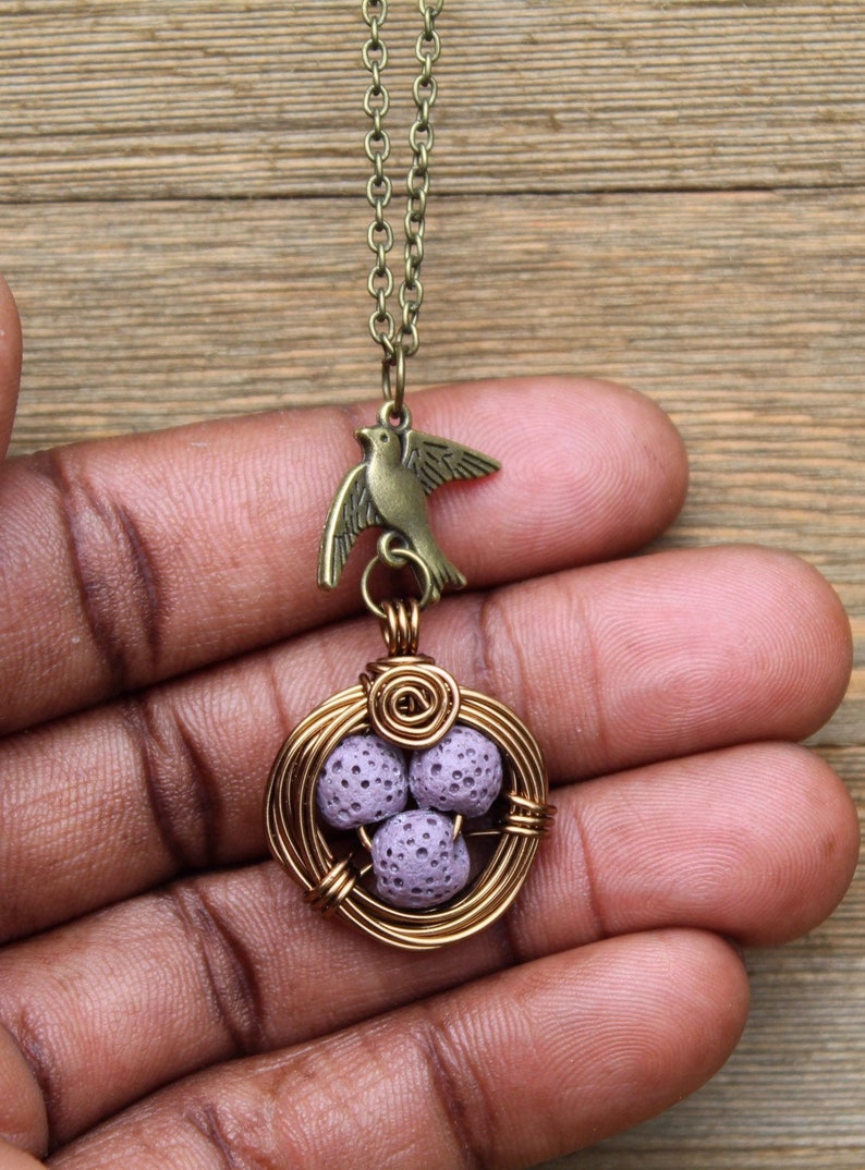 May include: A delicate necklace featuring a bird charm and a wire-wrapped nest with three lavender lava rock beads.