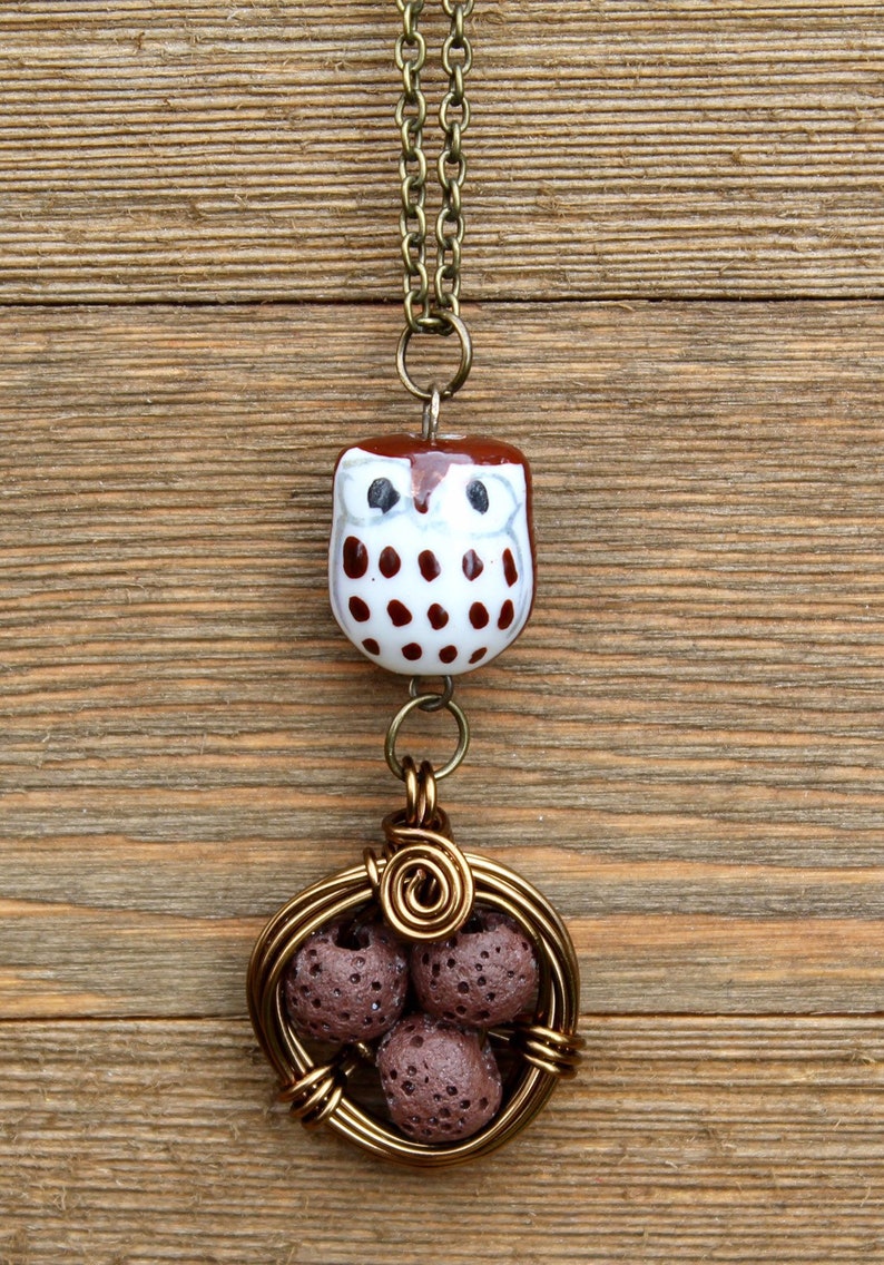 Puede incluir: Un dije de b&uacute;ho marr&oacute;n y blanco cuelga de una cadena dorada. El dije de b&uacute;ho est&aacute; unido a una jaula de alambre con cuatro cuentas de roca de lava marr&oacute;n en su interior.