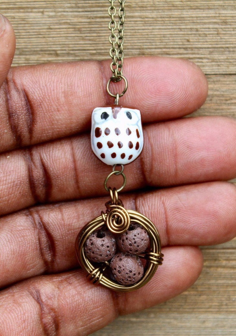 Puede incluir: Un collar de cadena dorada con un dije de b&uacute;ho marr&oacute;n y blanco y una jaula de alambre con cuentas de lava marr&oacute;n.