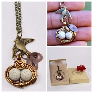 Puede incluir: Un collar de oro con un dije de pájaro, un nido con dos huevos blancos y dos dijes de cobre con los números 2 y 4. El collar se muestra sobre un fondo blanco.