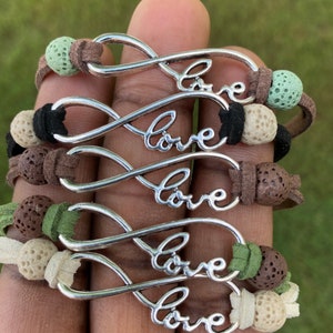 Puede incluir: Cinco pulseras con símbolo de infinito con la palabra "love" inscrita en cada una. Las pulseras están hechas de cordón de cuero marrón y tienen cuentas de roca volcánica en varios colores, incluyendo verde, negro y blanco.