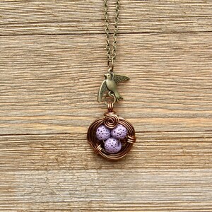 May include: A delicate necklace with a bird charm and a wire-wrapped nest with purple eggs. The necklace is on a wooden background.