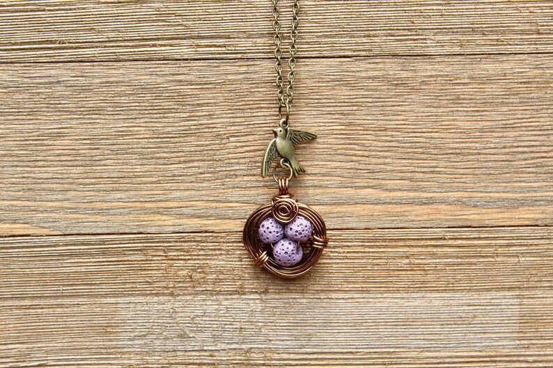 May include: A delicate necklace with a bird charm and a wire-wrapped nest with purple eggs. The necklace is on a wooden background.