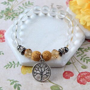 Op de afbeelding: Een armband van helder kwartskristal met bruine lavasteen kralen en een zilveren levensboom bedel. De armband ligt op een wit oppervlak met een bloemenpatroon.