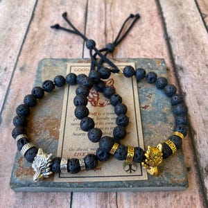 May include: Two black lava stone bracelets with silver and gold wolf head charms. The bracelets have a black cord closure and are on a slate background. The text on the card reads "Wolf is a symbol of loyalty and... hearts to control... Go ahead, we... LIVE YOUR... Spot"