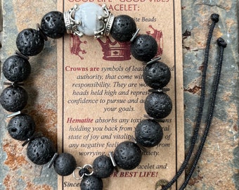 Op de afbeelding: Een armband van zwarte lavasteen kralen met een zilveren kroonbedel en een zwart koord. De armband ligt op een bruine achtergrond met tekst die "GOOD LIFE - GOOD VIBES" en "CROWNS ARE SYMBOLS OF LEADERSHIP, THAT COME WITH RESPONSIBILITY. THEY ARE WORN HEADS HELD HIGH AND REPRESENT CONFIDENCE TO PURSUE AND ACHIEVE YOUR GOALS." en "HEMATITE ABSORBS ANY TOXIC ENERGY HOLDING YOU BACK FROM YOUR STATE OF JOY AND VITALITY. IT ABSORBS ANY NEGATIVE FEELINGS, WORRYING, ANXIETY." en "SINCE SPOT TYSON" luidt.